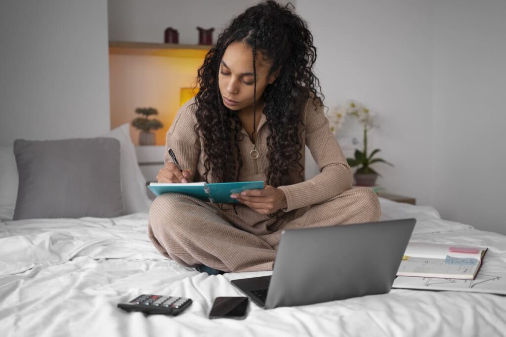 Mulher sentada na cama, escrevendo em um caderno enquanto usa um notebook, em um ambiente confortável de estudo.
