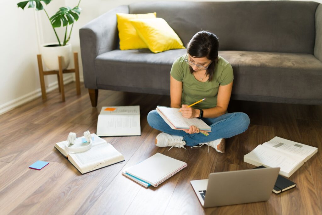 Jovem sentada no chão com as pernas cruzadas, estudando e escrevendo em um caderno, cercada por livros, materiais escolares e um notebook.