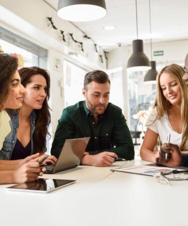 Grupo de profissionais reunido em uma mesa de trabalho, colaborando em uma reunião, com notebook, tablet e documentos.