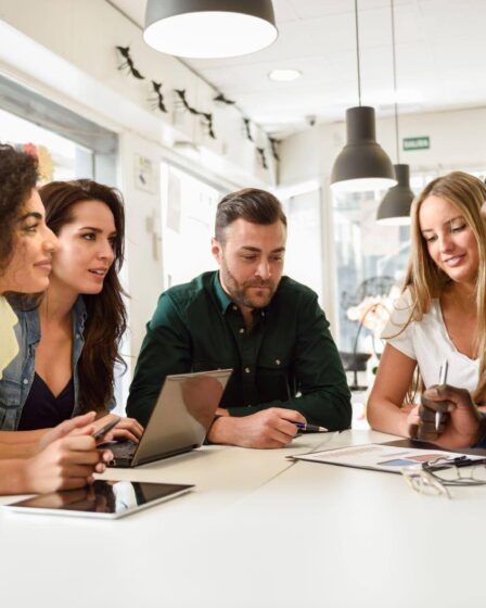 Grupo de profissionais reunido em uma mesa de trabalho, colaborando em uma reunião, com notebook, tablet e documentos.