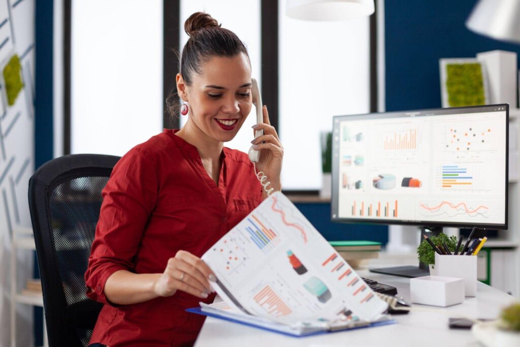 Mulher trabalha em um escritório analisando relatórios e gráficos enquanto fala ao telefone, ilustrando o perfil profissional analista com foco em interpretação de dados