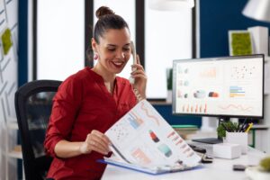 Mulher trabalha em um escritório analisando relatórios e gráficos enquanto fala ao telefone, ilustrando o perfil profissional analista com foco em interpretação de dados
