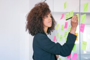 Mulher escrevendo em post-its coloridos fixados em um quadro de vidro, organizando metas com foco na aplicação da metodologia SMART.