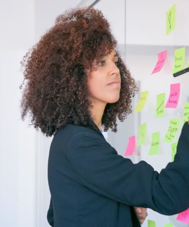 Mulher escrevendo em post-its coloridos fixados em um quadro de vidro, organizando metas com foco na aplicação da metodologia SMART.