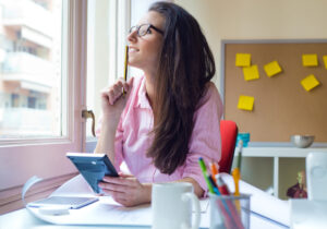 Mulher no escritório segurando uma caneta enquanto reflete sobre metas e prazos, com post-its ao fundo representando a aplicação prática da metodologia SMART.