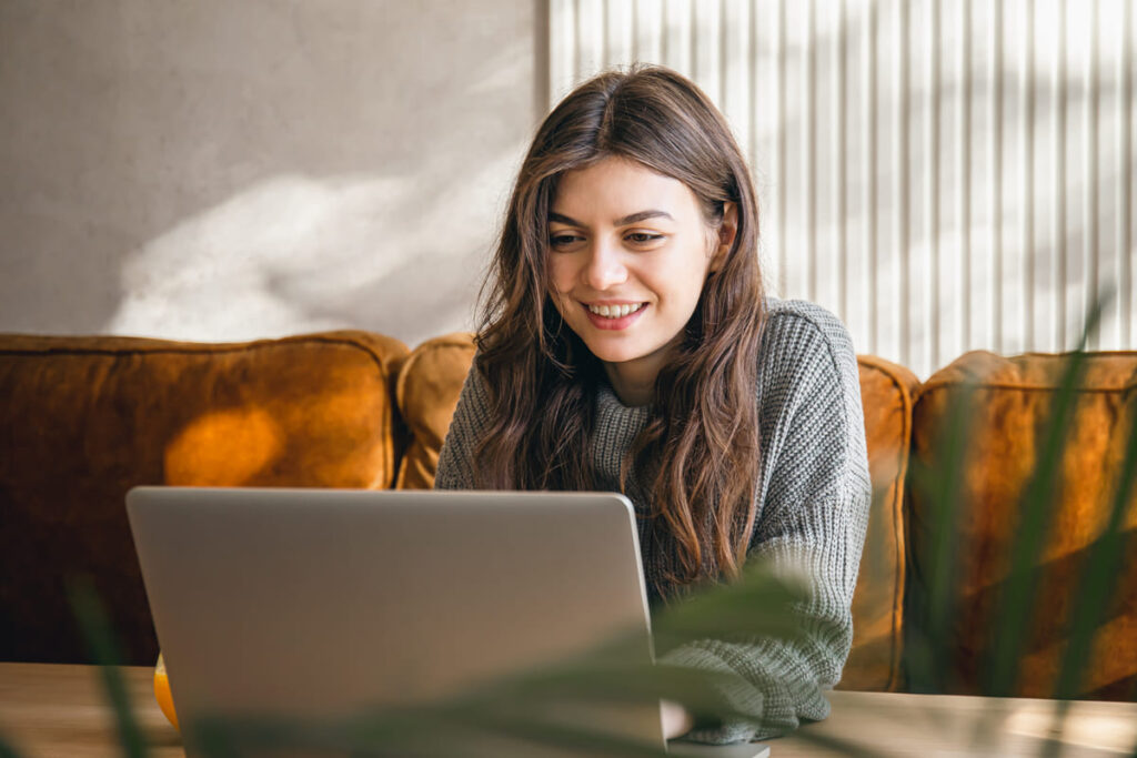 Estudante sorrindo enquanto acessa o notebook, sentada em um sofá na cor laranja.
