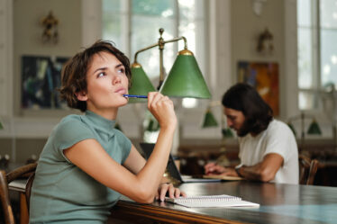 Estudante pensativa em uma biblioteca, segurando uma caneta e olhando para cima enquanto planeja seus estudos e pesquisa sobre o que é tecnólogo no ensino superior.
