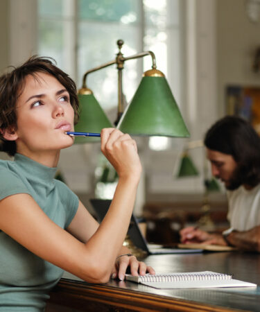 Estudante pensativa em uma biblioteca, segurando uma caneta e olhando para cima enquanto planeja seus estudos e pesquisa sobre o que é tecnólogo no ensino superior.