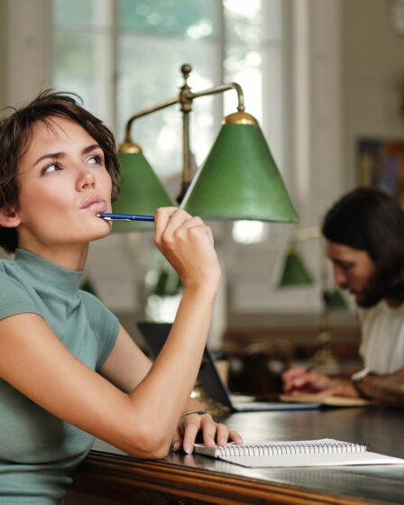 Estudante pensativa em uma biblioteca, segurando uma caneta e olhando para cima enquanto planeja seus estudos e pesquisa sobre o que é tecnólogo no ensino superior.