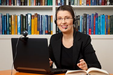 Professora sorridente com fone de ouvido, sentada à mesa em frente de um computador com uma estante de livros ao fundo.