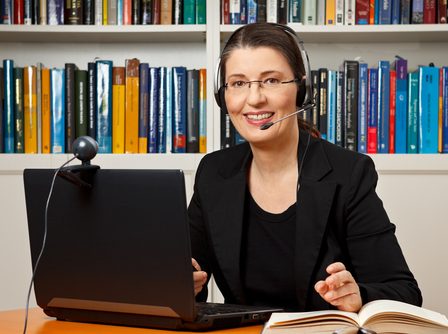 Professora sorridente com fone de ouvido, sentada à mesa em frente de um computador com uma estante de livros ao fundo.