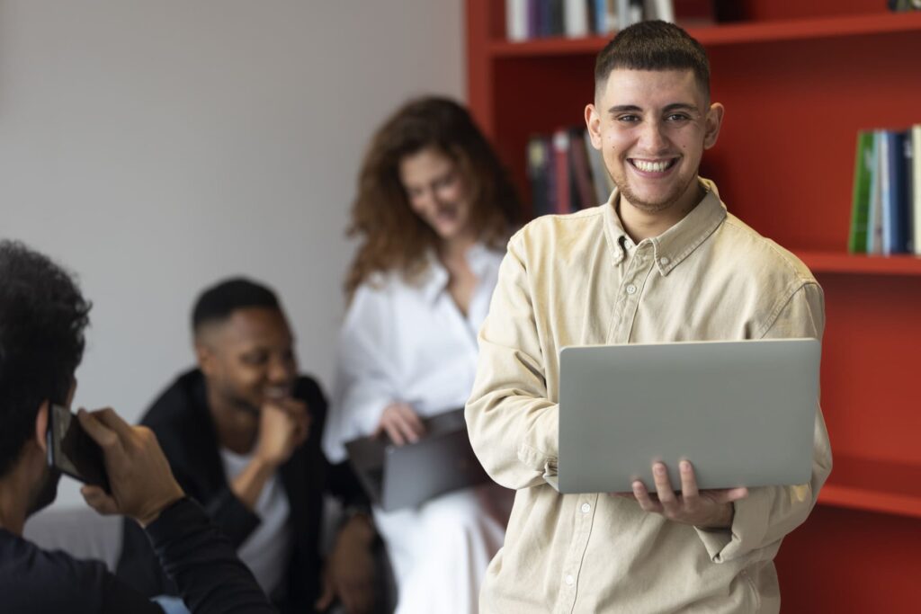 Jovem sorrindo enquanto usa um notebook em escritório, representando oportunidades de estágio e trainee em ambiente corporativo.