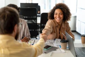 Jovem profissional celebra oportunidade de primeiro emprego ao cumprimentar recrutador durante reunião em ambiente de trabalho.