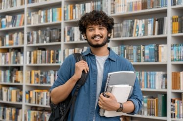 Jovem estudante do sexo masculino segurando livros e com mochila nas costas sorri diante de uma estante de livros. Ref: 2071252046