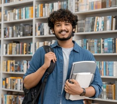 Jovem estudante do sexo masculino segurando livros e com mochila nas costas sorri diante de uma estante de livros. Ref: 2071252046