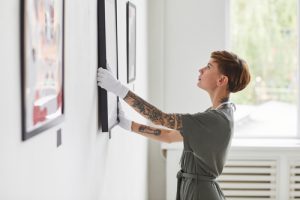 Jovem formada em Artes Visuais, com braços tatuados, de cabelos curtos e usando luvas, pendura quadro em galeria de arte.