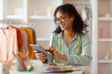 Empreendedora jovem, com semblante feliz, utilizando um tablet e envolta a fitas métricas e papéis com fontes coloridas em uma loja de roupas em construção ao fundo.