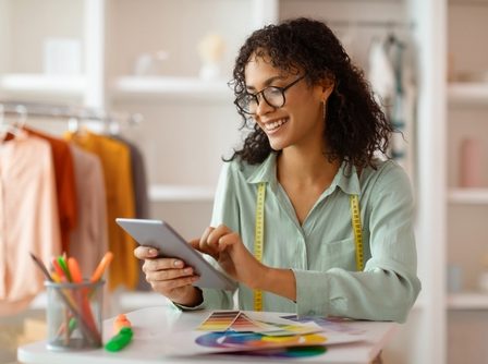 Empreendedora jovem, com semblante feliz, utilizando um tablet e envolta a fitas métricas e papéis com fontes coloridas em uma loja de roupas em construção ao fundo.
