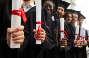 Grupo de graduandos de beca e diploma em mãos, enfileirados e felizes no dia da colação de grau.