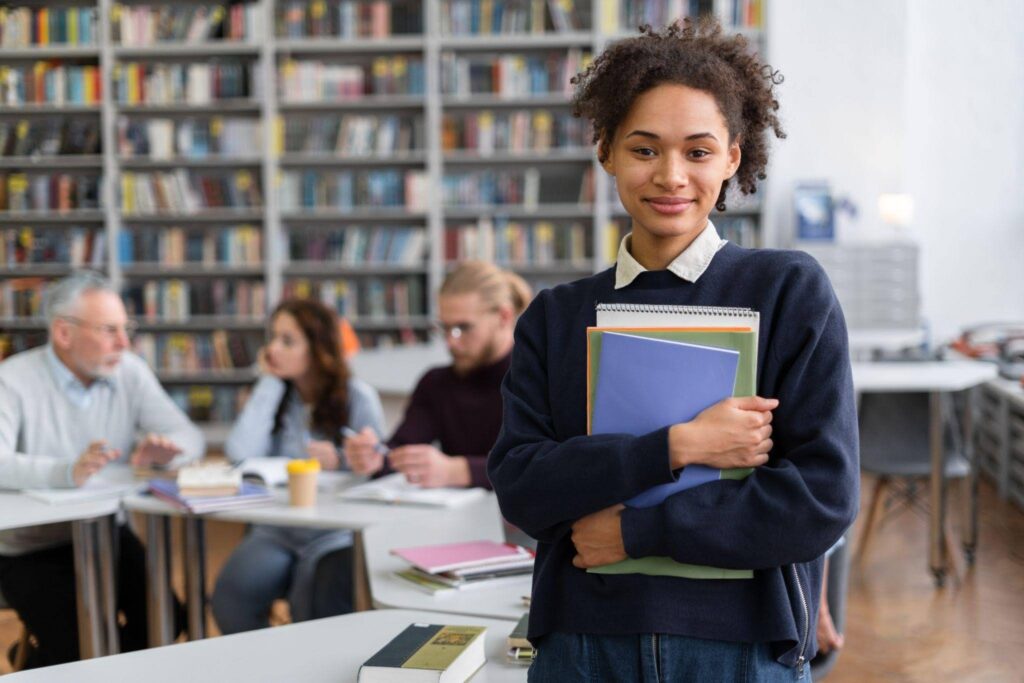Estudante sorrindo em uma biblioteca, segurando livros e cadernos, com pessoas estudando ao fundo.