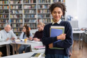 Estudante sorrindo em uma biblioteca, segurando livros e cadernos, com pessoas estudando ao fundo.