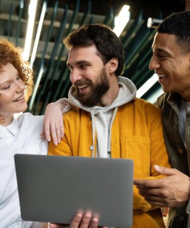 Três colegas sorrindo com notebook, representando jovens no mercado de trabalho.