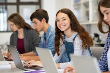 Quatro jovens sentados em frente de seus notebooks, três mulheres e um homem, com uma das meninas olhando para a câmera com semblante feliz e animado e os outros sérios encarando a tela do computador.