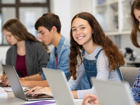 Quatro jovens sentados em frente de seus notebooks, três mulheres e um homem, com uma das meninas olhando para a câmera com semblante feliz e animado e os outros sérios encarando a tela do computador.