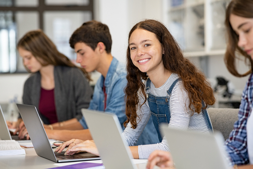 Quatro jovens sentados em frente de seus notebooks, três mulheres e um homem, com uma das meninas olhando para a câmera com semblante feliz e animado e os outros sérios encarando a tela do computador.