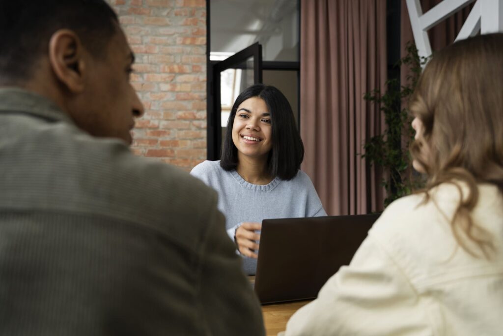 Grupo de três pessoas conversando em escritório moderno, com uma mulher sorrindo enquanto fala durante uma entrevista coletiva de emprego