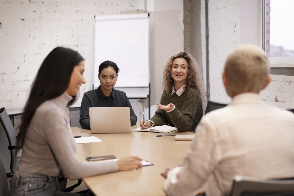 Equipe reunida em mesa de reunião com laptops e anotações, enquanto uma mulher apresenta ideias em uma entrevista coletiva de emprego em ambiente profissional