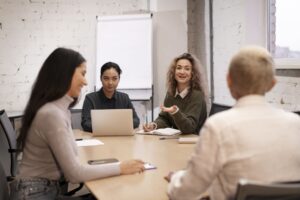 Equipe reunida em mesa de reunião com laptops e anotações, enquanto uma mulher apresenta ideias em uma entrevista coletiva de emprego em ambiente profissional