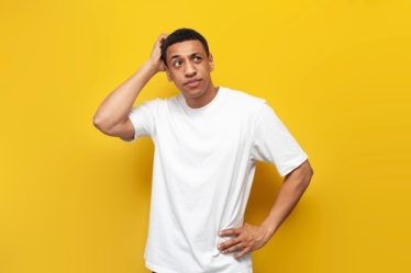 jovem de camiseta branca em fundo amarelo, com a mão na cabeça expressando dúvida.