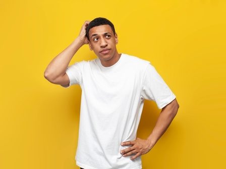 jovem de camiseta branca em fundo amarelo, com a mão na cabeça expressando dúvida.