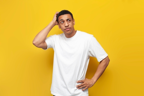 jovem de camiseta branca em fundo amarelo, com a mão na cabeça expressando dúvida.