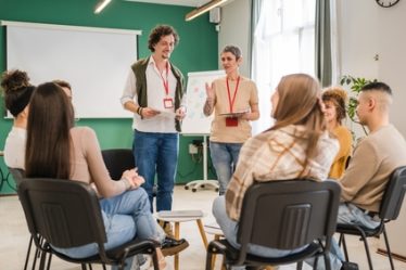 Imagem de um grupo de pessoas sentadas em círculo, ouvindo orientações de dois recrutadores.