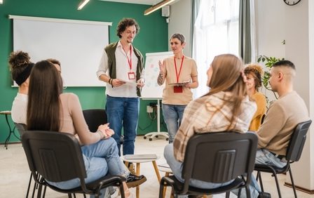 Imagem de um grupo de pessoas sentadas em círculo, ouvindo orientações de dois recrutadores.