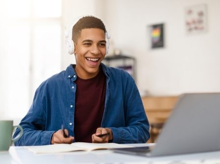 Estudante com semblante feliz, em frente a uma mesa com computador, fones de ouvido, um caderno e com canetas nas mãos.