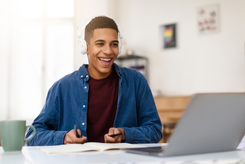 Estudante com semblante feliz, em frente a uma mesa com computador, fones de ouvido, um caderno e com canetas nas mãos.