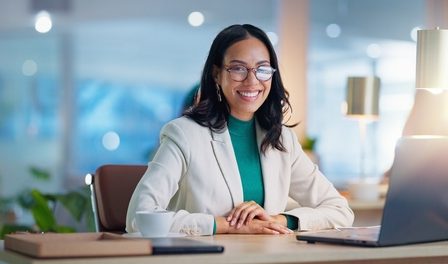 Empreendedora jovem, com semblante feliz, sentada em sua mesa, em frente a um notebook com luminária e artigos de escritório em sua mesa.