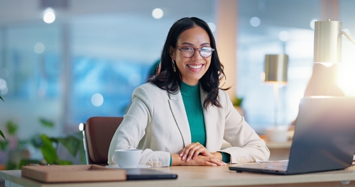 Empreendedora jovem, com semblante feliz, sentada em sua mesa, em frente a um notebook com luminária e artigos de escritório em sua mesa.