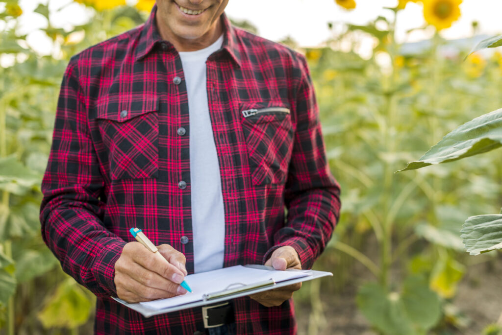 Homem sorrindo em plantação, enquanto anota em prancheta, representando a carreira em Agronomia no campo.