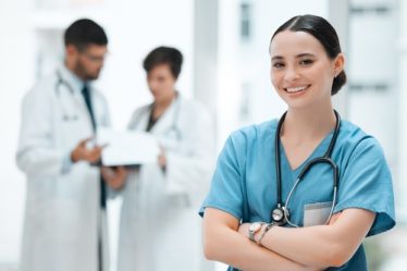 Enfermeira de braços cruzados, usando uniforme azul e estetoscópio, sorrindo em um ambiente hospitalar