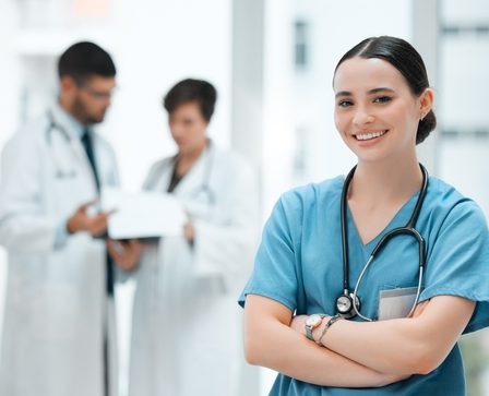 Enfermeira de braços cruzados, usando uniforme azul e estetoscópio, sorrindo em um ambiente hospitalar