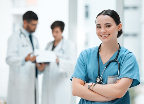 Enfermeira de braços cruzados, usando uniforme azul e estetoscópio, sorrindo em um ambiente hospitalar