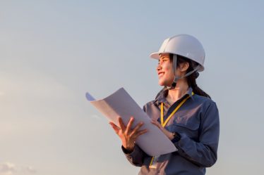 Jovem engenheira mulher usando capacete de segurança, com um uniforme de trabalho e cinto de ferramentas, em um ambiente de construção ou obra, olhando para o horizonte com um sorriso confiante.