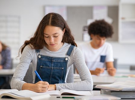 Estudante sentada na carteira realizando uma prova com o uso de uma caneta.