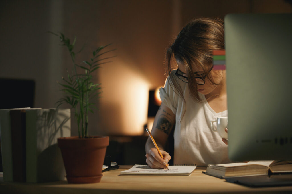 Mulher usando óculos, sentada à mesa e escrevendo em um caderno enquanto estuda.