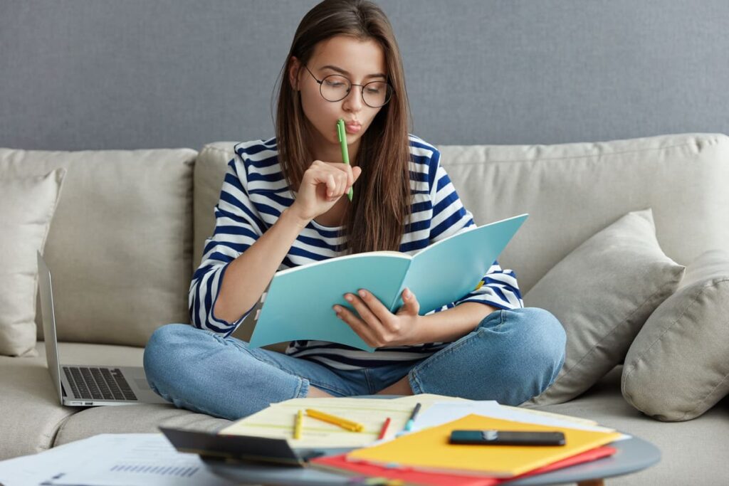 Mulher de óculos sentada em um sofá, estudando com um caderno aberto e segurando uma caneta próxima à boca.