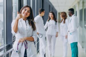 Jovem médica em corredor de hospital segurando um estetoscópio, com outros profissionais ao fundo, ilustrando profissões na área da saúde.